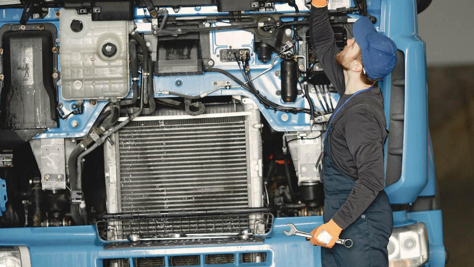 Mecânico com boné e macacão azul inspecionando o motor de um caminhão azul, com o capô levantado e segurando uma chave inglesa.